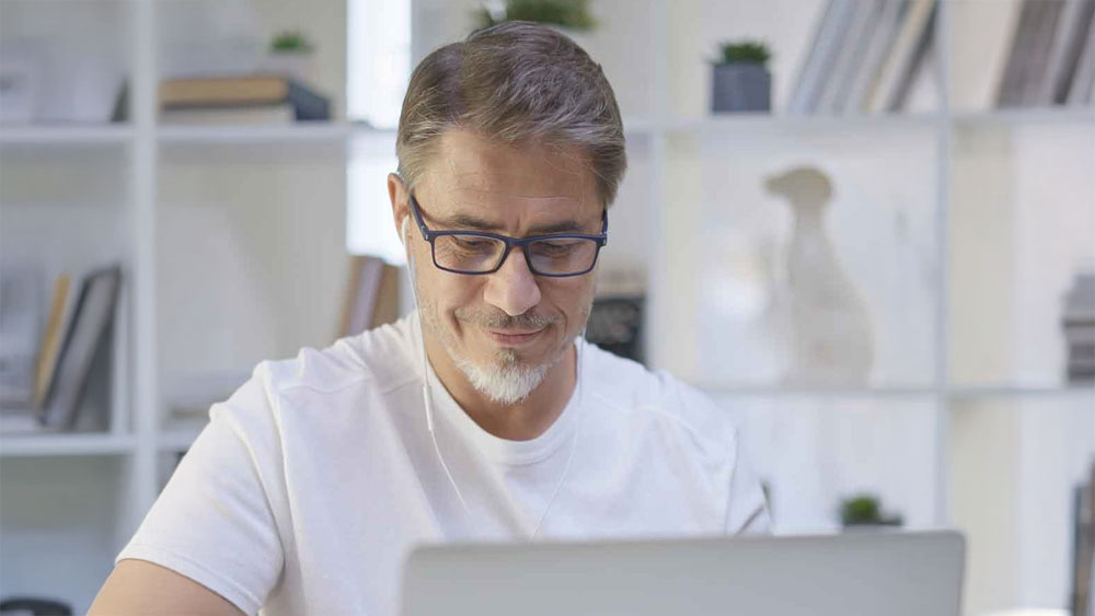 A middle-aged man with glasses and a short beard, wearing a white t-shirt, smiles while working on a laptop in a bright room, perhaps handling Reese Technologies Hardware Maintenance, with shelves and books in the background.