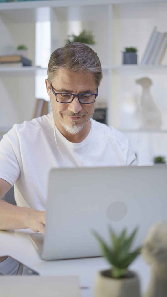 A middle-aged man with glasses and earphones sits at a desk, smiling while using a laptop. Shelves with books and small plants are in the background, as he researches About Reese Technologies, with a potted plant on the desk in the foreground.