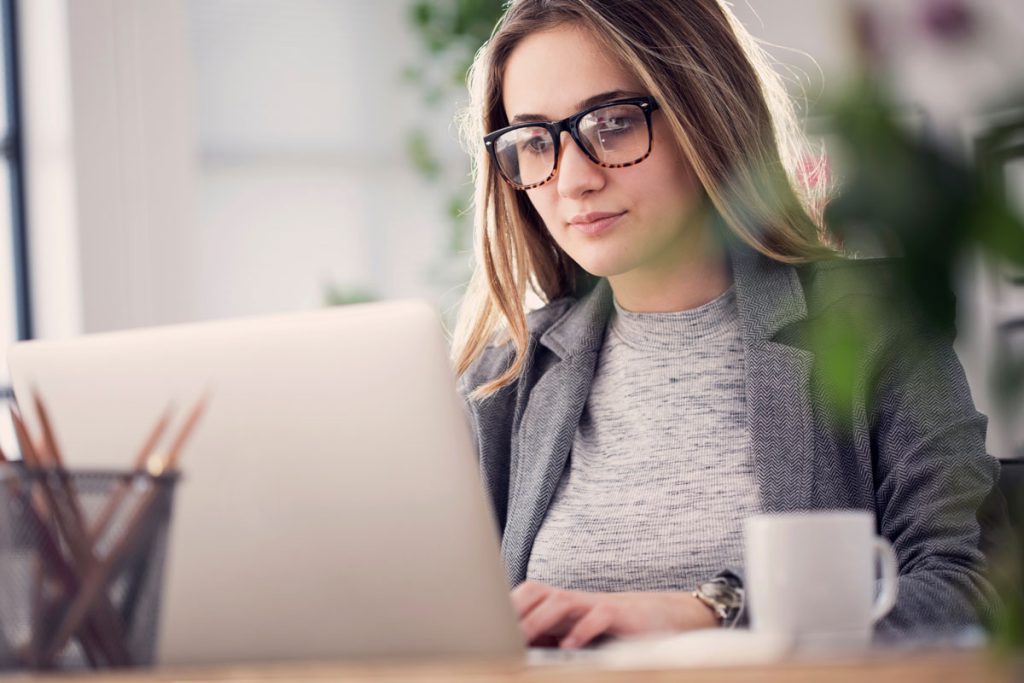 A woman wearing glasses and a gray blazer works on a laptop at a desk. There is a white coffee mug and a pencil holder with pencils nearby. She appears focused and is indoors, with plants in the background.