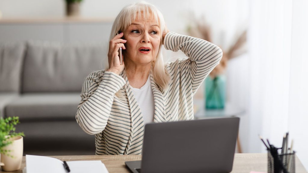An older woman with long gray hair sits at a desk with a laptop, holding a phone to her ear and touching her head, looking worried or stressed. Papers and plants are on the desk in a bright room.
