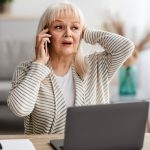 An older woman with long gray hair sits at a desk with a laptop, holding a phone to her ear and touching her head, looking worried or stressed. Papers and plants are on the desk in a bright room.