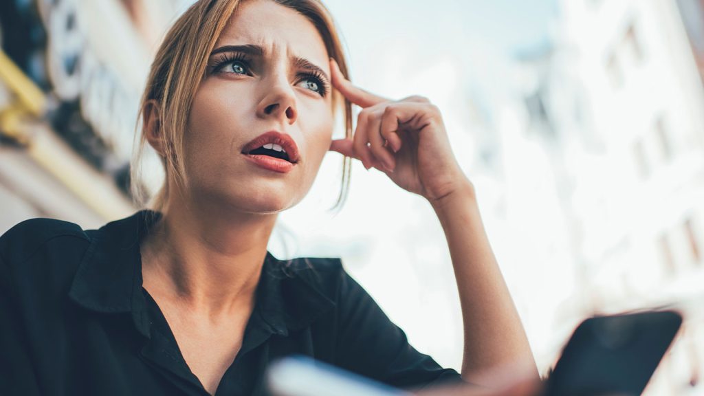 A young woman with light brown hair and a black shirt sits outdoors, looking confused or concerned. She holds her head with one hand and a smartphone with the other, with a blurred city background behind her.