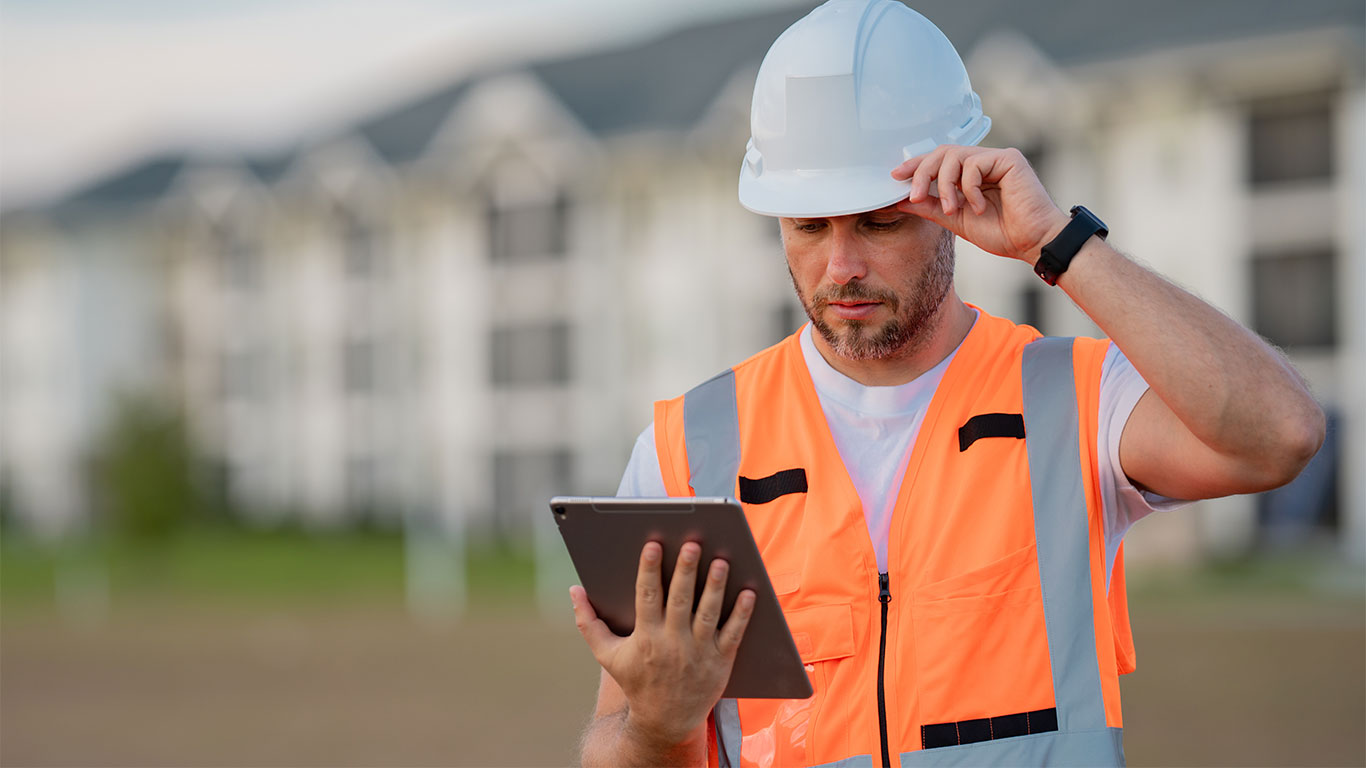 A construction worker wearing a white hard hat and orange safety vest looks at a tablet outdoors, with blurred buildings in the background.