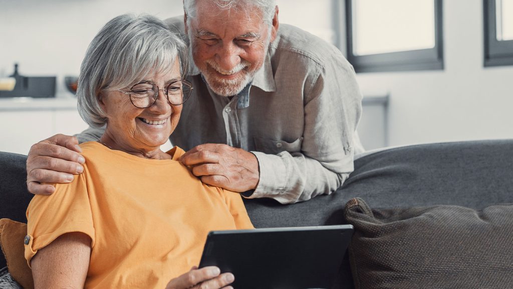 An older woman wearing glasses and a yellow shirt sits on a couch holding a tablet, smiling, while an older man stands close behind her, smiling and resting his hand on her shoulder.