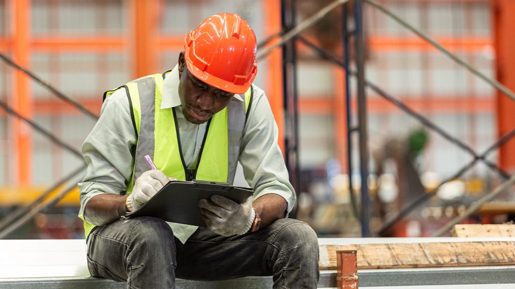 A construction worker wearing a safety vest, gloves, and an orange hard hat sits and writes on a clipboard or tablet in a warehouse, leveraging Microsoft 365 to handle employee disputes efficiently in the industrial setting.