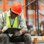 A construction worker wearing a safety vest, gloves, and an orange hard hat sits and writes on a clipboard or tablet in a warehouse, leveraging Microsoft 365 to handle employee disputes efficiently in the industrial setting.