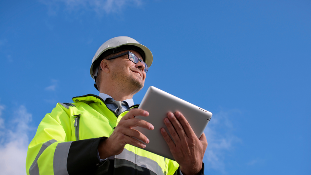 A person wearing a hard hat, safety glasses, and a bright yellow safety jacket holds a tablet outdoors, looking upward against a blue sky with a few clouds.