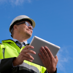 A person wearing a hard hat, safety glasses, and a bright yellow safety jacket holds a tablet outdoors, looking upward against a blue sky with a few clouds.