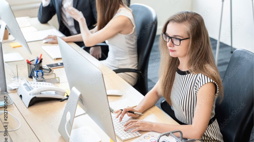A woman wearing glasses and a striped dress works at a computer—choosing the right office monitor for efficiency—in a modern, collaborative workspace where others are also focused at their desks.