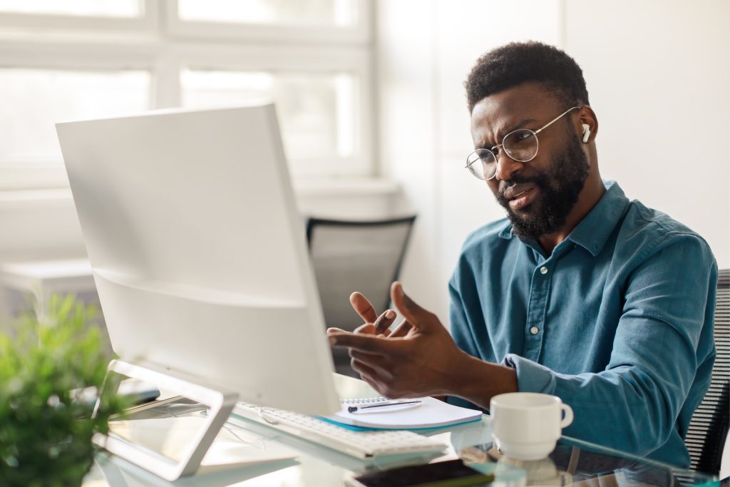 A man wearing glasses and earbuds sits at a desk in front of a computer, looking confused as he gestures with one hand—possibly struggling with choosing the right office monitor. A notebook, keyboard, and coffee cup are on the desk.