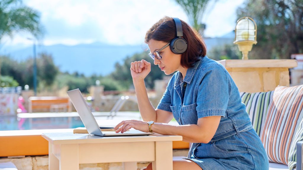 A woman wearing headphones and a denim dress sits outside by a pool, working on her laptop at a small table—perhaps researching, "Is That Email From a Hacker? Here's How to Tell." Cushions, a lamp, trees, and mountains complete the serene scene.