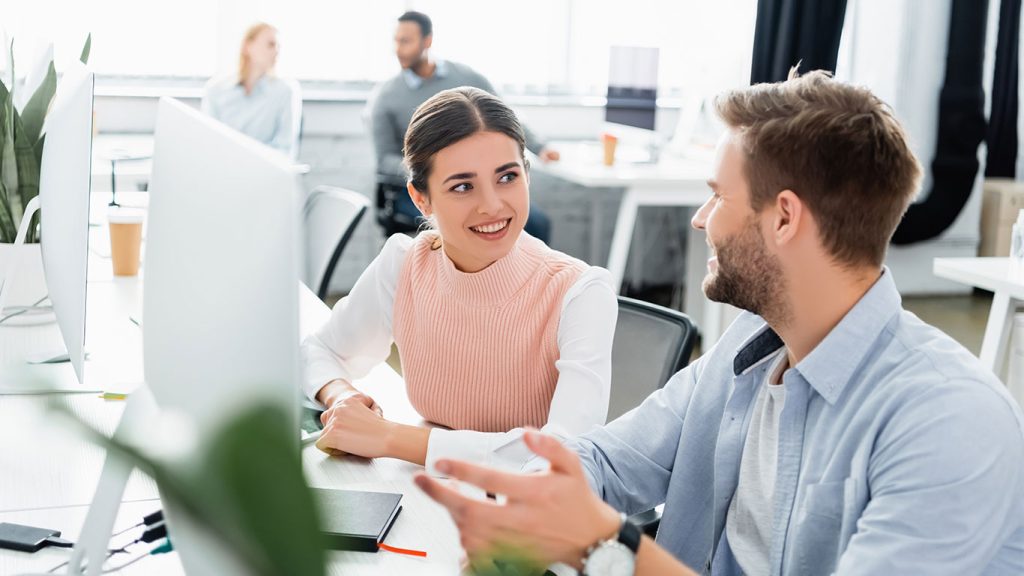 Two colleagues sit at a desk in a modern office, smiling and discussing how to set up Microsoft 365 Apps on your new phone. The environment appears collaborative and bright, with computers, plants, and coworkers working in the background.