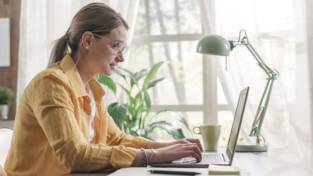 A woman wearing glasses and a yellow shirt types on a laptop at a desk by a window. A green desk lamp, coffee mug, and plants are visible, creating a bright, cozy workspace.